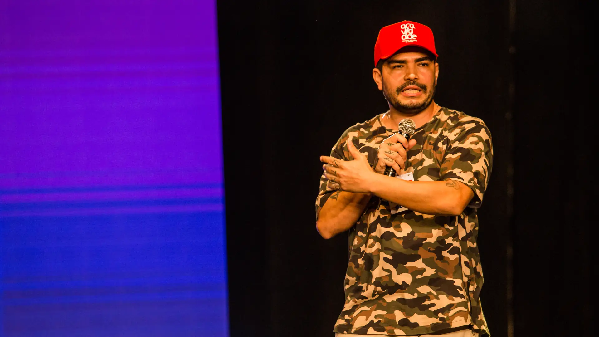 Ticko Bboy ministrando a palestra O Marketing do Artista no palco, de boné vermelho e camiseta camuflada, em Votuporanga